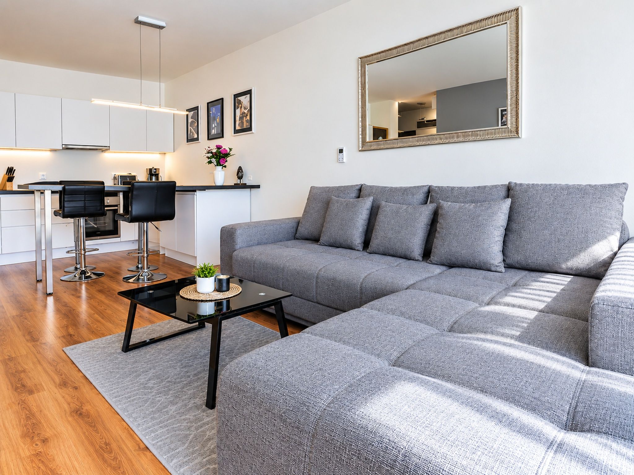 Ein modernes Wohnzimmer mit einem grauen Ecksofa, einem Couchtisch und einer Küchenzeile im Hintergrund.