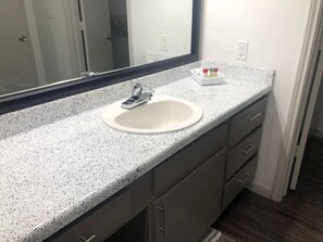 Een badkamer met een wit beklede wastafel, een witte wasbak, een spiegel en een klein dienblad met toiletartikelen.