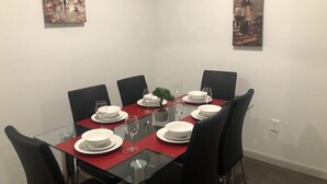 Un coin repas avec une table en verre dressée pour six personnes, entourée de chaises noires. Deux tableaux ornent les murs, une petite plante en pot est posée sur la table et un set de table rouge se trouve sous chaque assiette.