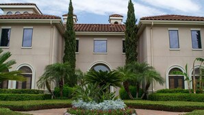 Ein Gebäude mit einer symmetrischen Fassade, Bogenfenstern und einem zentralen, kreisförmigen Garten mit Palmen und Sträuchern.