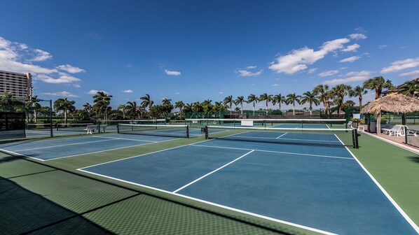 Un complexe de courts de tennis comprenant plusieurs terrains, des palmiers et un pavillon à toit de chaume.