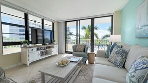A modern living room with a flat-screen TV, a sofa, a coffee table, and a balcony with a view of palm trees and buildings. - South Seas T4- 306: Beautiful end unit condo, Beach Front (Marco Island)