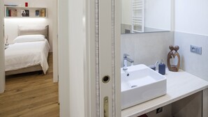 Un dormitorio con cama, mesita de noche con libros y lámpara, y un baño con lavamanos, espejo y toallero.