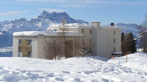 Un bâtiment moderne dont le toit est recouvert de neige, entouré d’un paysage enneigé et avec des montagnes en arrière-plan.
