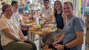 Un groupe de personnes est assis à une table à l’intérieur d’un restaurant doté de grandes fenêtres et de plantes suspendues.