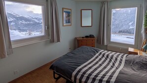 Une chambre à coucher avec un lit, deux fenêtres donnant sur un paysage de montagne enneigé, une commode en bois et un ventilateur de plafond.