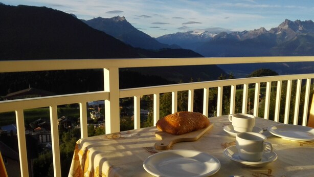 Une table avec des assiettes, des tasses et un pain, placée sur un balcon donnant sur un paysage montagneux.