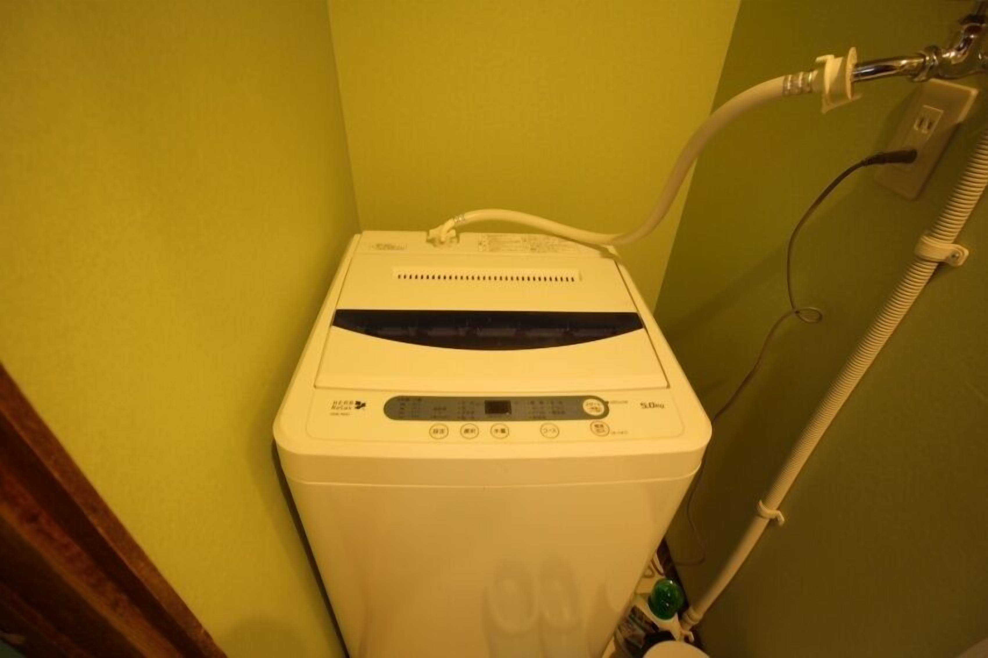 A compact, white washing machine with a control panel, connected to a hose.