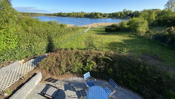 Eine Terrasse mit Blick auf einen See, umgeben von Grünpflanzen und einem Heckenstreifen.
