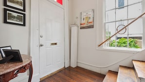 Een kamer met een houten vloer, een witte deur, een tafeltje met een fotolijstje en een raam met uitzicht op groen.