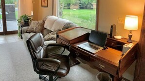 Een werkkamer met een laptop op een bureau, een lederen stoel, een bank en een groot raam met uitzicht op groen.