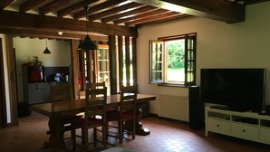 A cozy dining area with a wooden table and chairs, a hanging light fixture, a window with a view of greenery, and a television on a stand. - Charming traditional Norman House near Honfleur & Deauville  (Cormeilles)