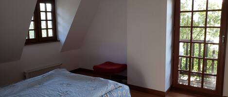 A bedroom with a sloped ceiling, a radiator, a bed with a floral bedspread, a red chair, and a large window with a view of greenery.