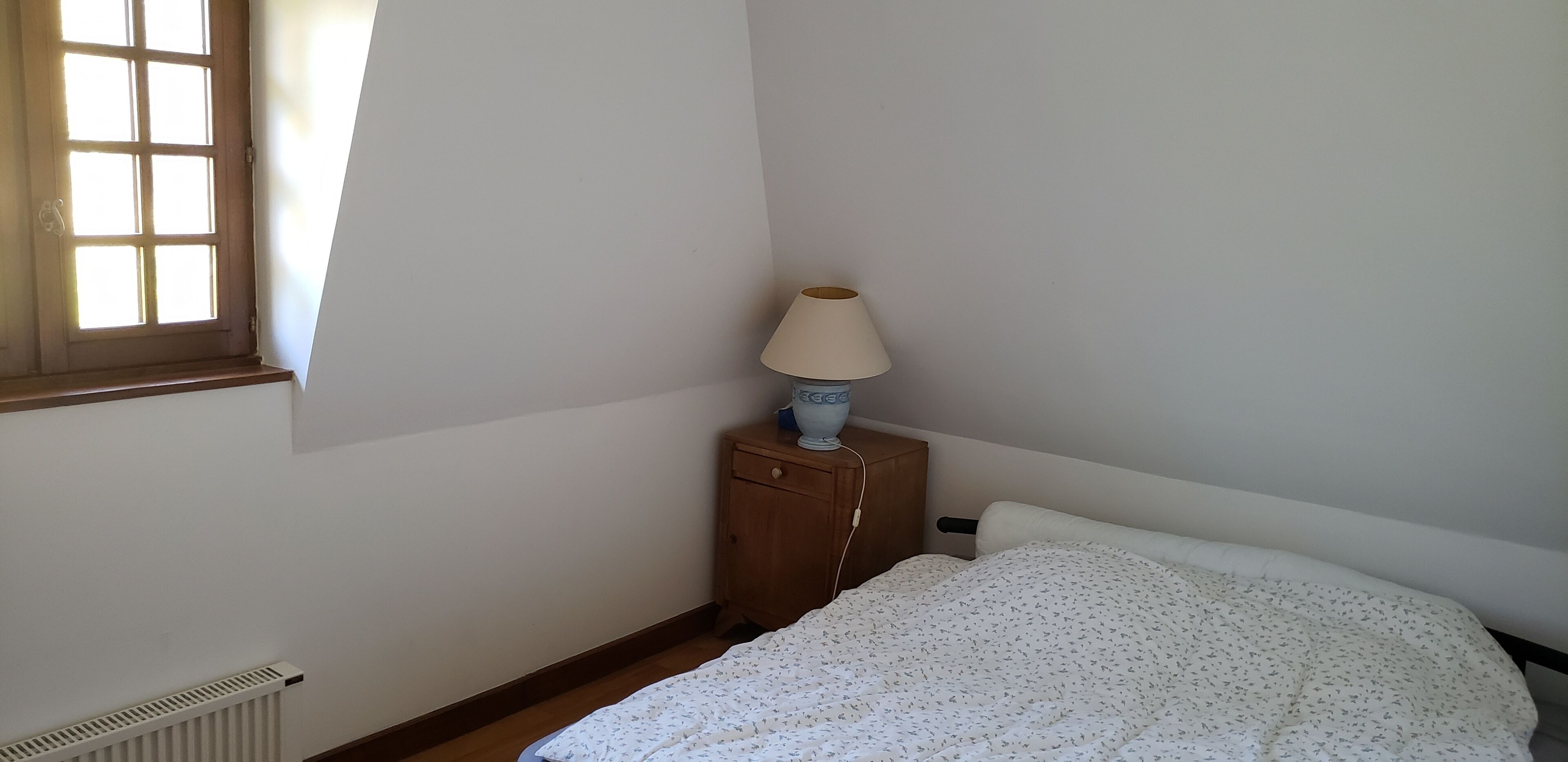 Ein Schlafzimmer mit Bett, Nachttisch mit Lampe, Heizkörper und Fenster.