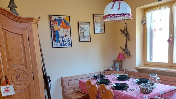 Ein Essbereich mit einem Holztisch, Stühlen und einer Bank. An der Wand hängen Bilder in Rahmen, eine Uhr und ein Fenster mit Vorhängen.