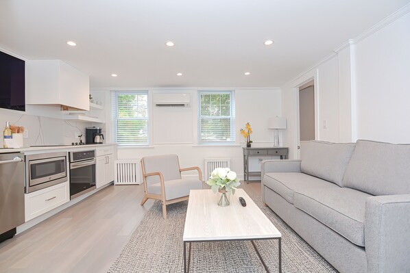 Una cocina moderna con electrodomésticos integrados, un comedor con mesa y sillas blancas, y un sofá gris.