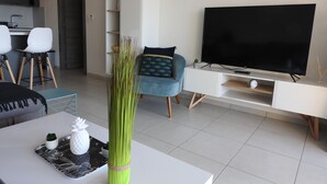 Ett modernt vardagsrum med en platt-TV, ett matbord dekorerat med en ananas, en hög grön växt och en blå soffa.