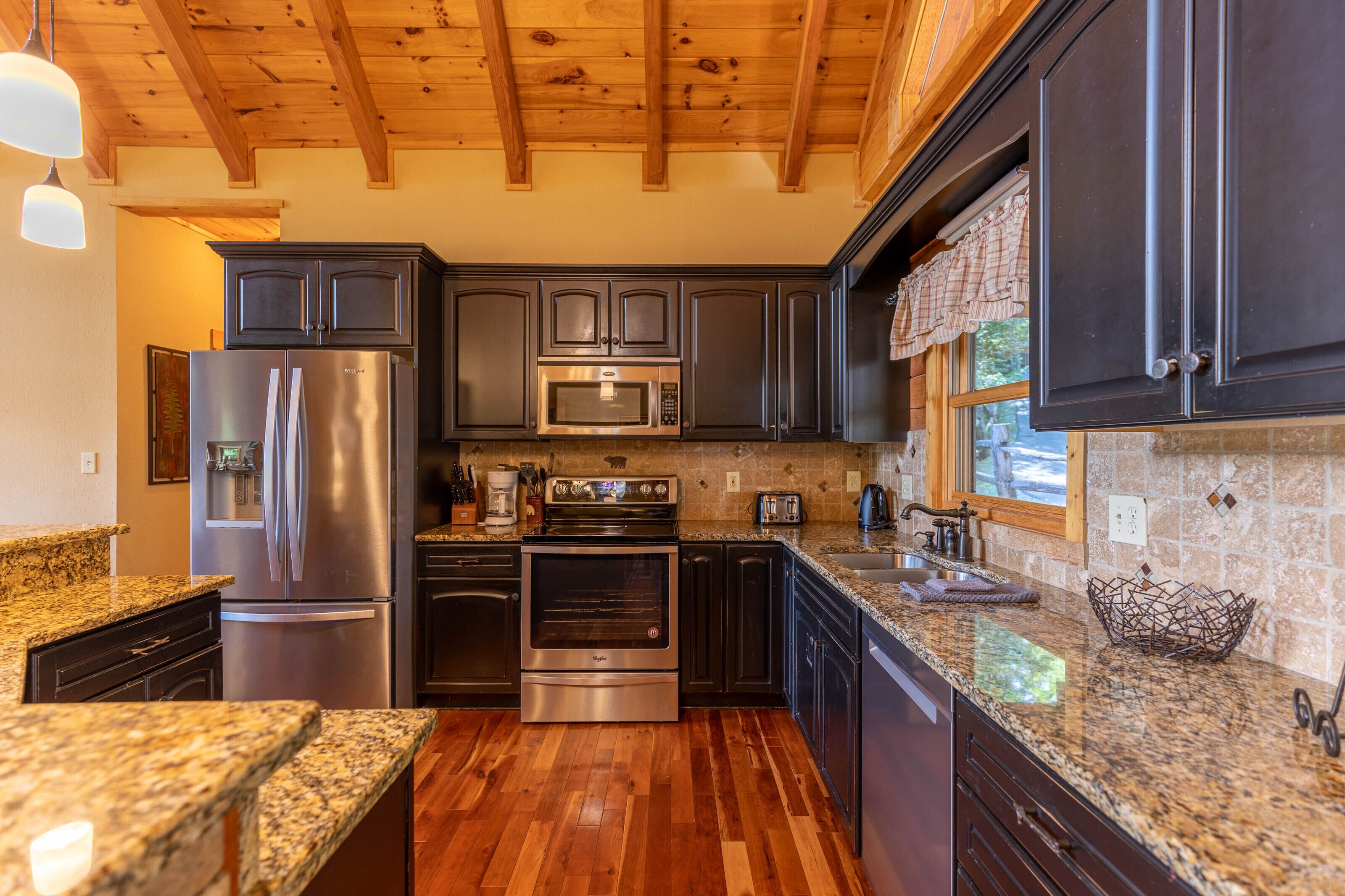 A modern kitchen with dark cabinetry, stainless steel appliances, granite countertops, and a wooden ceiling.