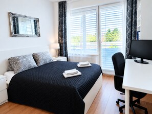 A hotel room with a bed, a desk with a computer, a chair, a window with blinds, and a mirror on the wall. - E2 Luxury OldTown Apt~2xFree Parking~Supermarket~PCScreen~Crib~Aircon~FastWifi (Košický kraj)
