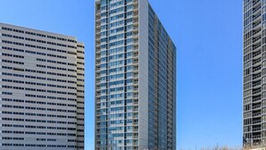 A modern high-rise building with multiple floors and balconies, surrounded by a sandy area with a wooden fence.