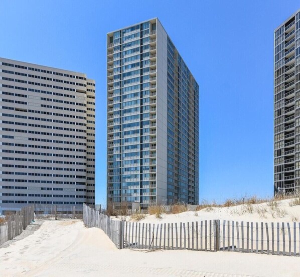 Un immeuble moderne de grande hauteur, doté de nombreux étages et de balcons, entouré d’une zone sablonneue délimitée par une clôture en bois.