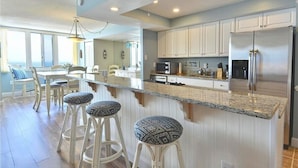 A modern kitchen with white cabinetry, a stainless steel refrigerator, and a granite countertop. There are three bar stools with patterned cushions.