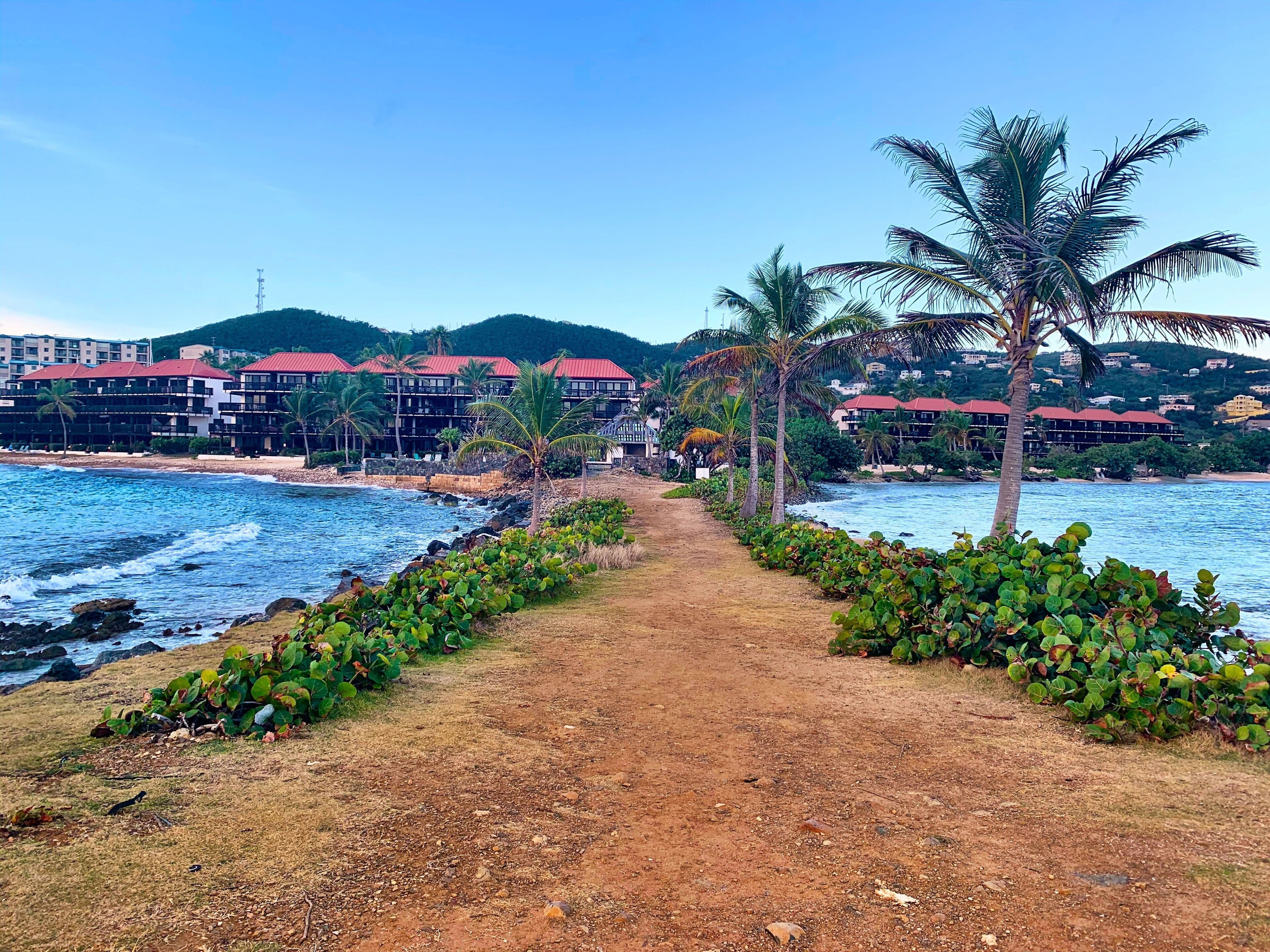 A coastal path leading to a beachfront hotel with red-roofed buildings, palm trees, and a clear view of the ocean.