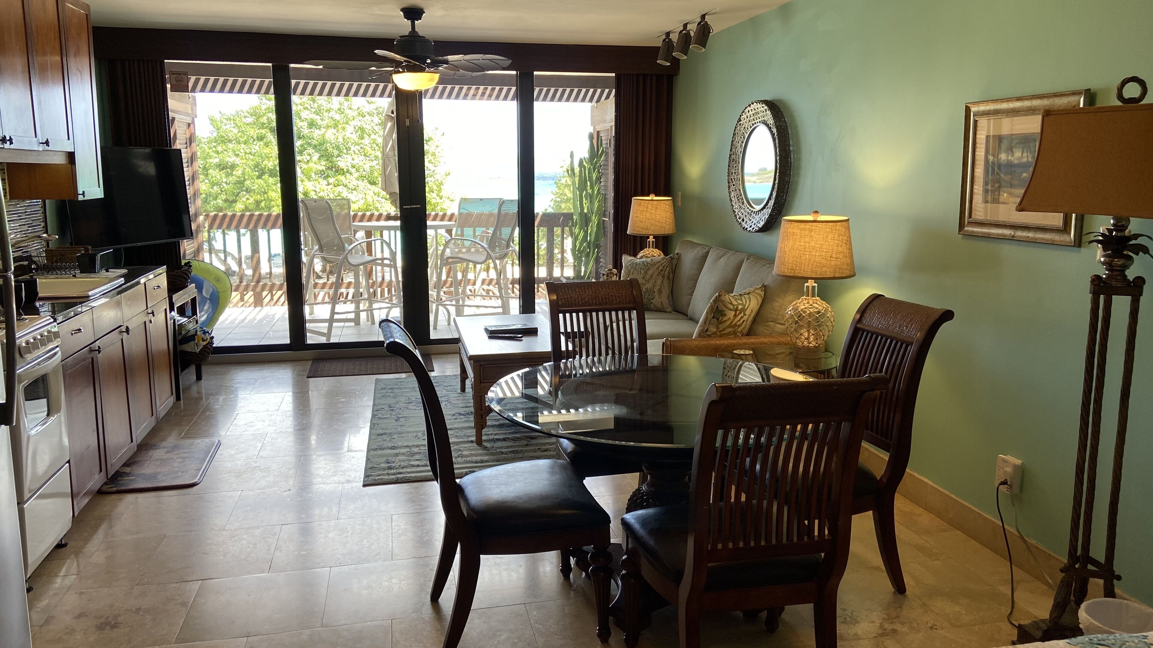 A dining area with a glass table, wooden chairs, and a sofa with cushions. There is a ceiling fan, a lamp, and a view of the outdoors through large windows.
