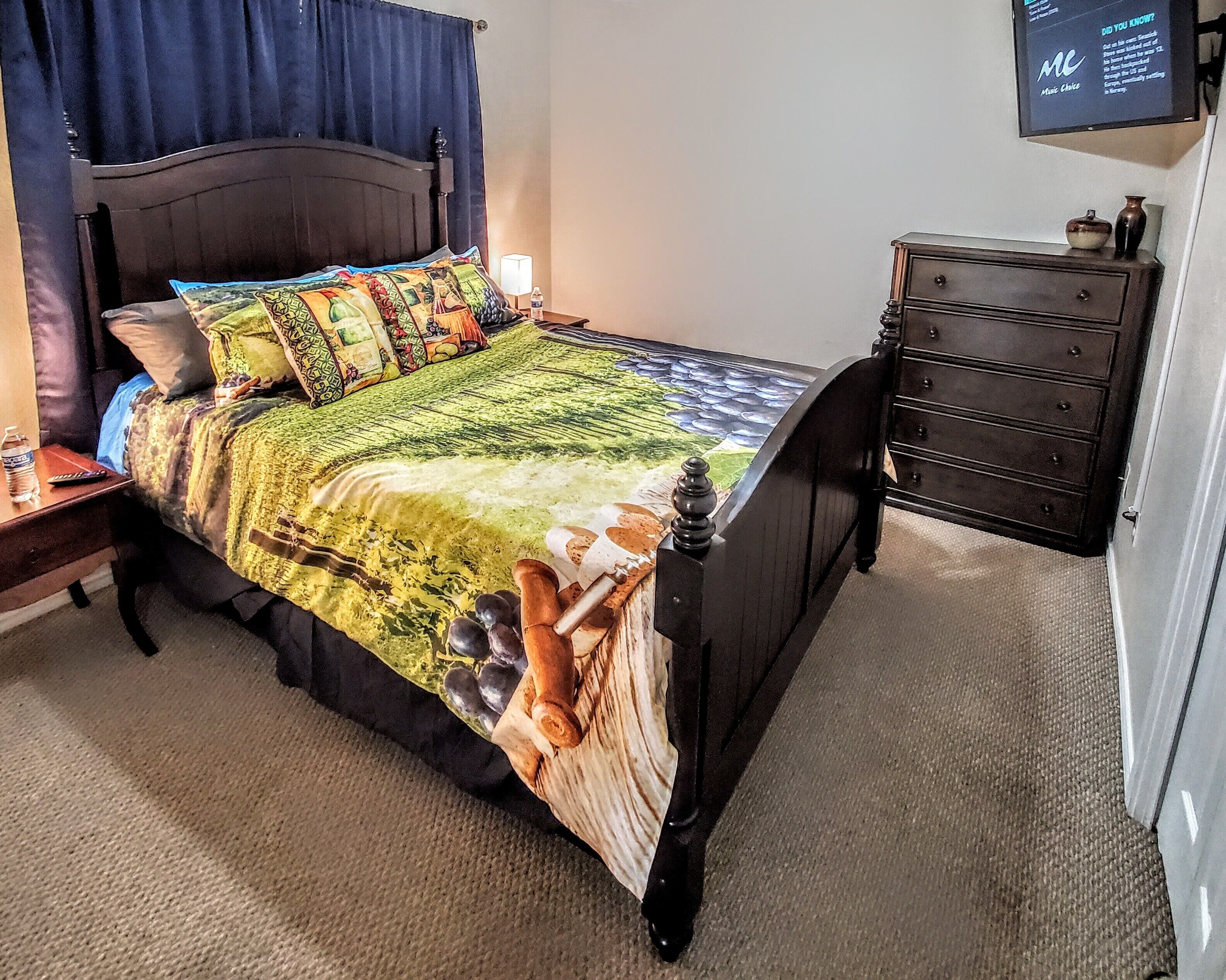 A neatly made bed with a colorful quilt, a bedside table with a bottle and remote control, and a dark wooden dresser with a TV on the wall.