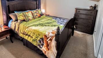 A neatly made bed with a colorful quilt, a bedside table with a bottle and remote control, and a dark wooden dresser with a TV on the wall.
