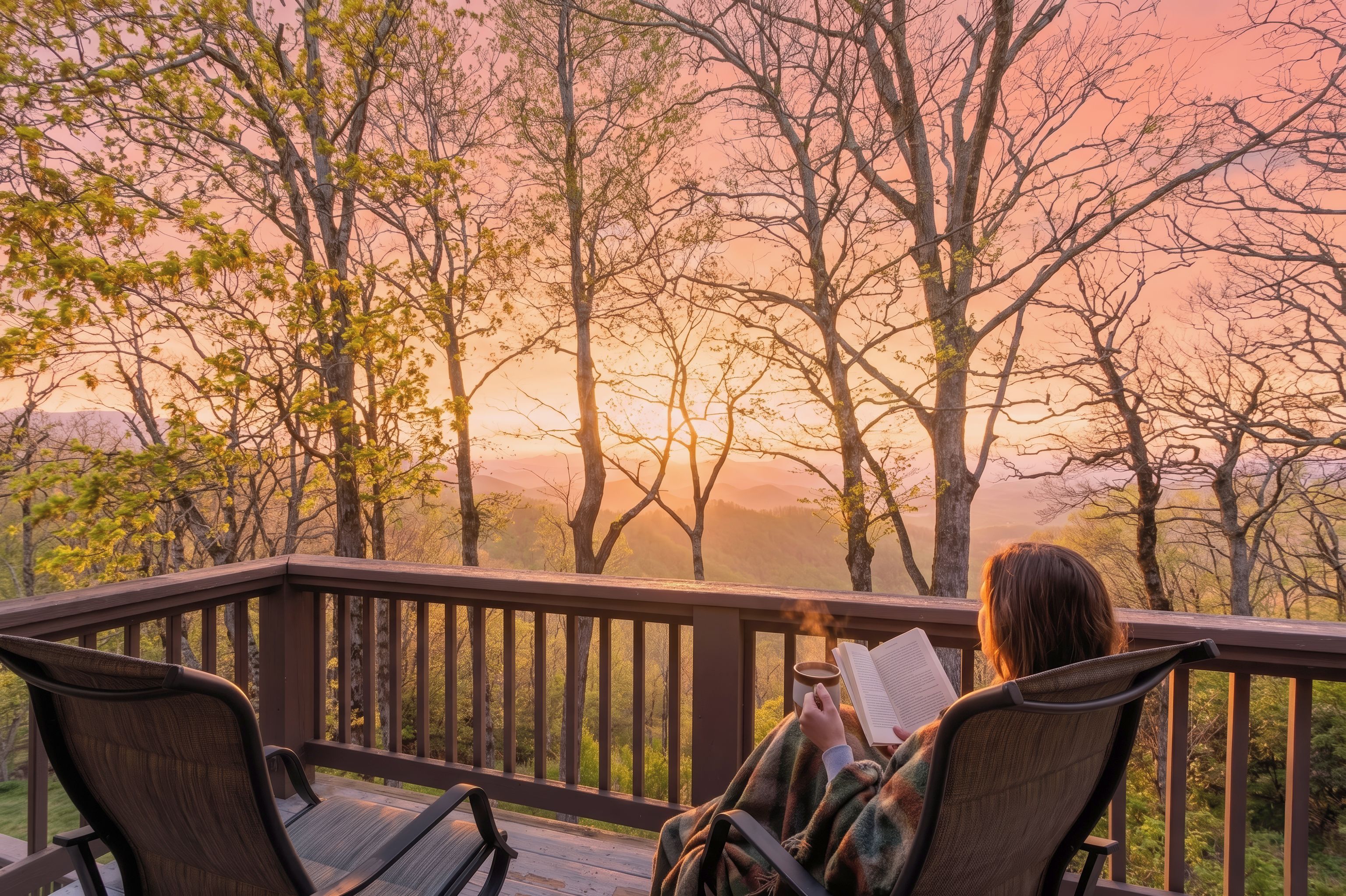 A person reading on a deck with a view of trees and a sunset.