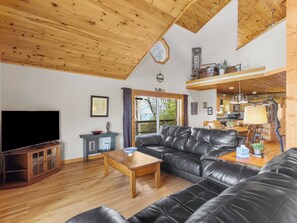Een woonkamer met een houten plafond, een zwarte leren bank, een houten salontafel, een televisie en uitzicht op buiten.
