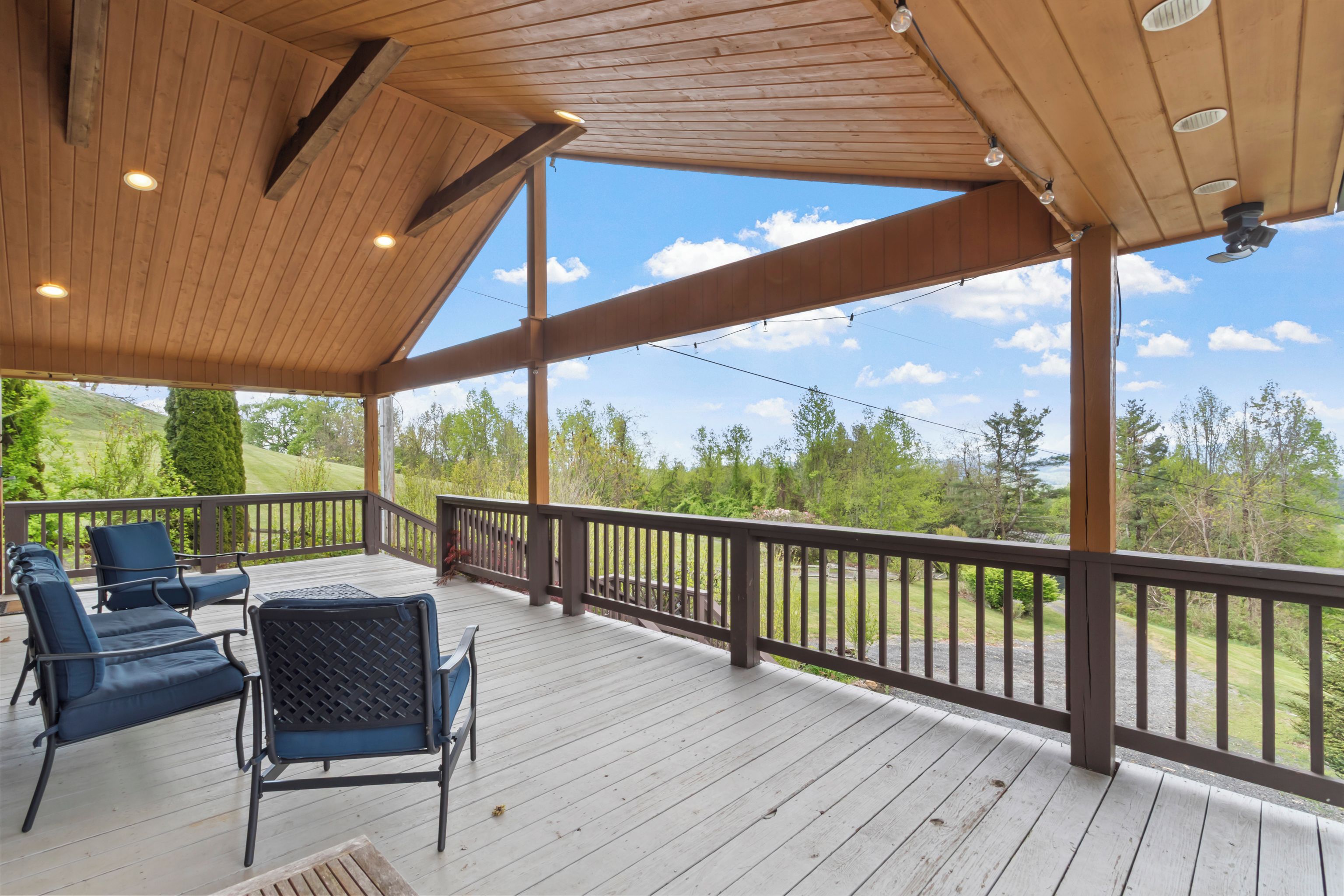 A covered outdoor deck with wooden railings, a seating area with chairs, and a view of a green landscape.