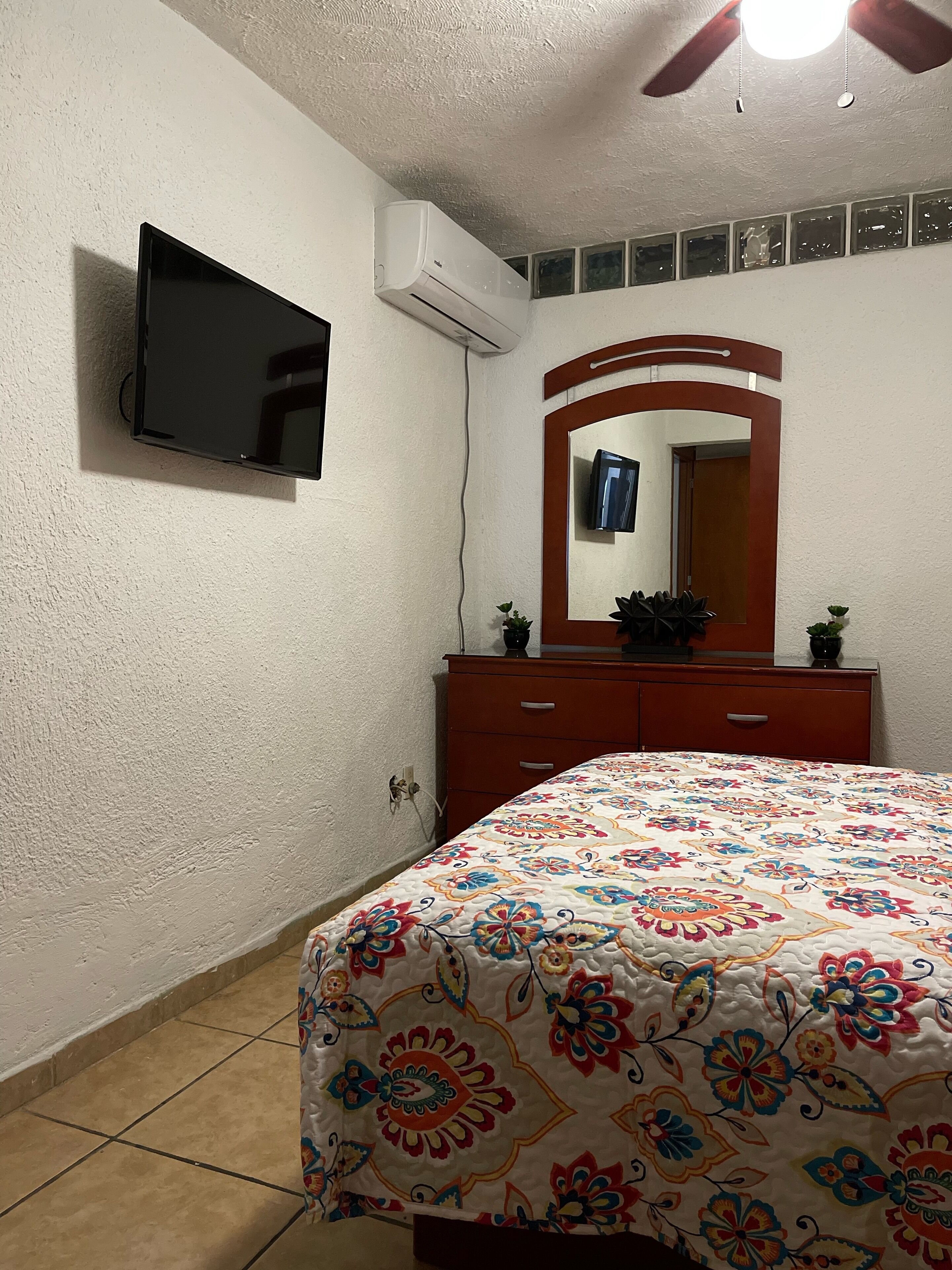 Une chambre à coucher comprenant un lit, un téléviseur fixé au mur, un climatiseur, une commode, un miroir et un ventilateur de plafond.