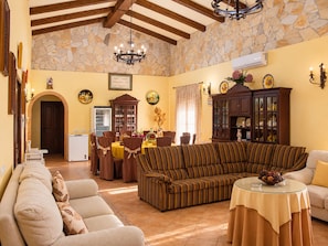 A spacious living room with a large striped sofa, a round coffee table, and a dining area with a wooden cabinet and a refrigerator. - Casa con Wifi e Impresionantes Vistas en Casabermeja (Casabermeja)
