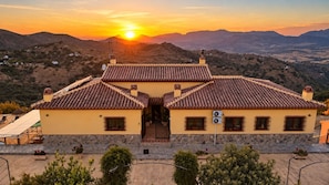 A two-story building with a tiled roof, a stone base, and a view of the surrounding landscape during sunset. - Casa con Wifi e Impresionantes Vistas en Casabermeja (Casabermeja)