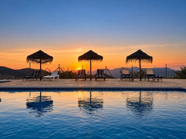 A poolside area with lounge chairs and umbrellas, surrounded by a fence and mountains in the background.