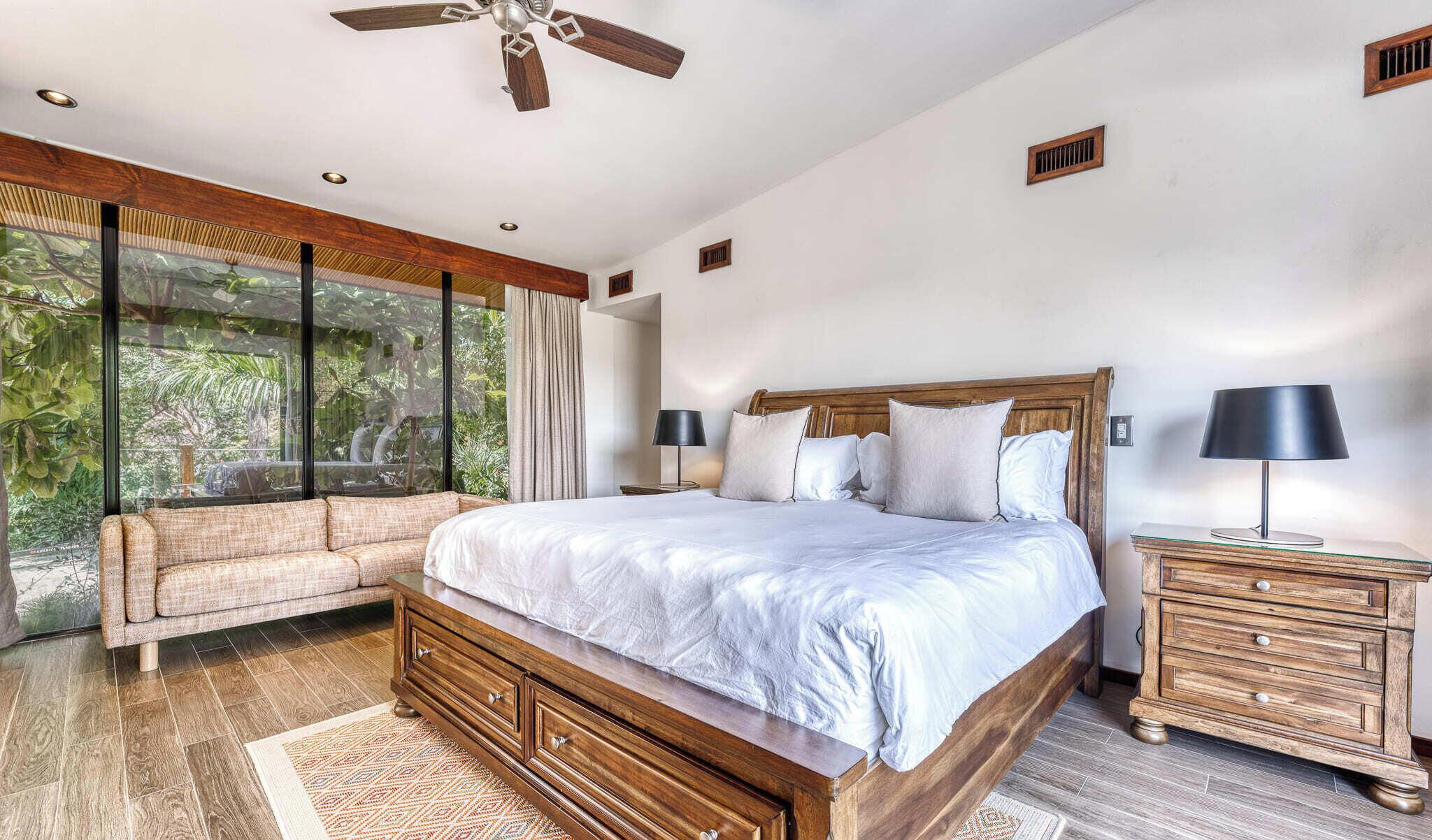 A bedroom with a large bed, a wooden nightstand, a sofa, and a ceiling fan.