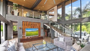 Amplia sala de estar con televisor grande, asientos cómodos, una escalera y vistas al exterior.