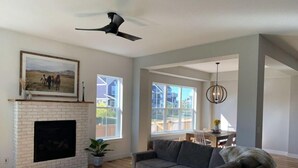 Amplia sala de estar con chimenea, un sofá grande de color gris y un comedor con luz natural proveniente de ventanas grandes.