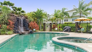 Piscina com cachoeira, rodeada por plantas tropicais e cerca.