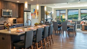 A modern kitchen with a central island, dark wood cabinets, stainless steel appliances, and a dining area with a large window offering a view of the outdoors.