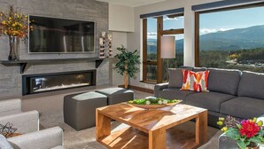 A modern living room with a flat-screen TV, fireplace, comfortable seating, and large windows offering a mountain view.