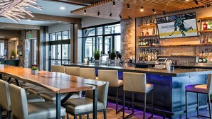 A modern bar area with a long wooden table and several bar stools. There is a large screen displaying a hockey game, and shelves stocked with bottles and glasses.