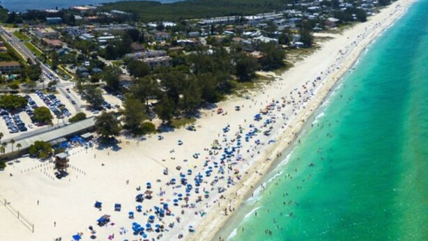 An aerial view of a beach with a long stretch of sand, a clear turquoise sea, and a dense residential area with houses and parked cars. - Beautifully furnished Condo (Bradenton)