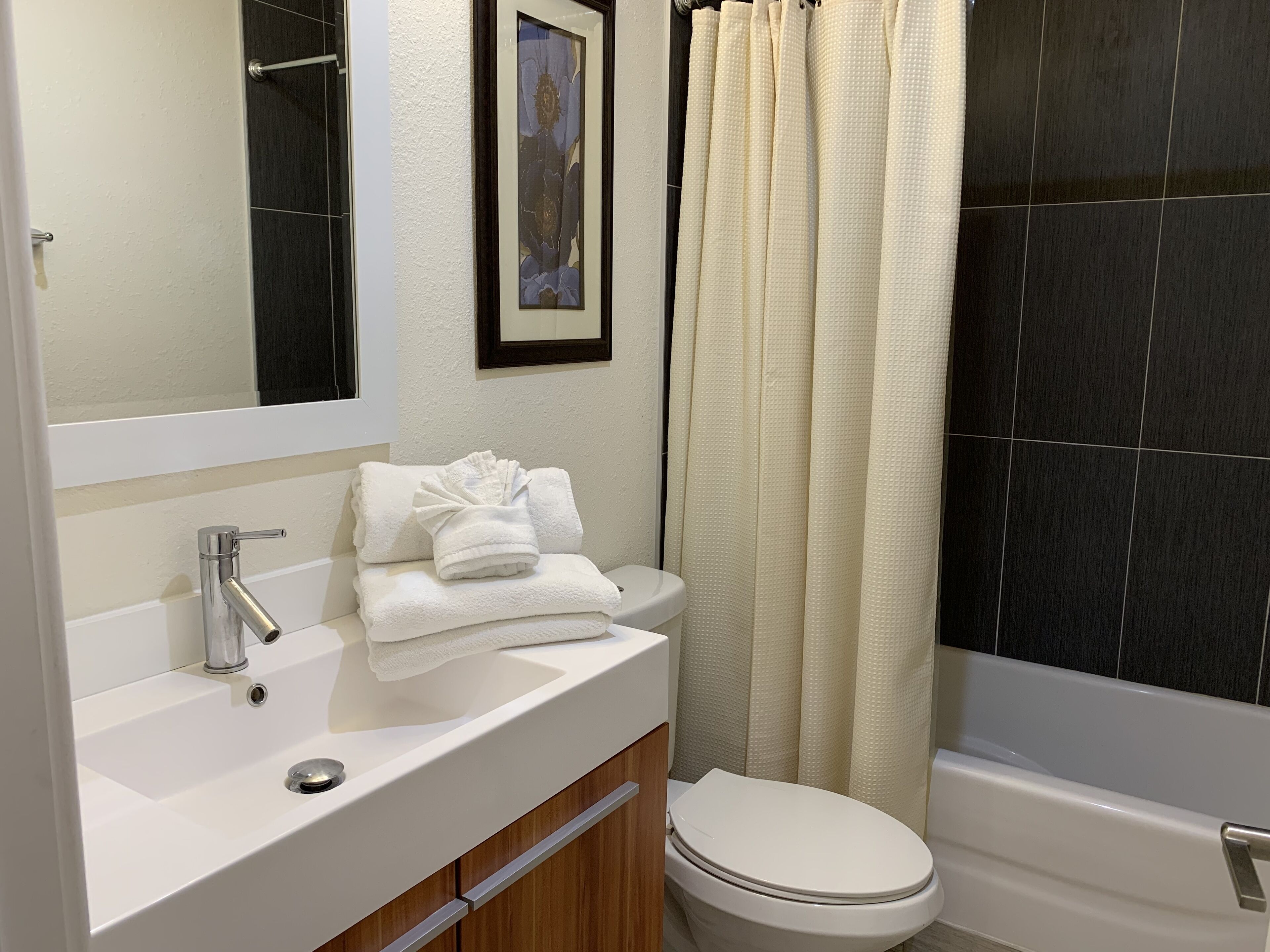 A bathroom with a white sink, a framed picture on the wall, a toilet, and a bathtub with a beige shower curtain.