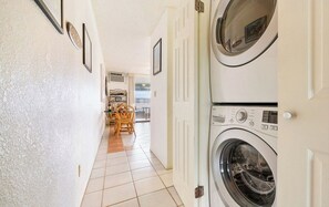 A laundry room with two stacked washing machines, a dining area with wicker chairs, and a hallway leading to a balcony. - Seaside Kona Condo/Top Fl, Sunsets, Private Lanai,Best Kona Location, Sleeps 2 (Kailua-Kona)