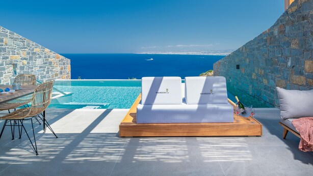 A sunlit outdoor seating area with a stone wall, a pool, and a view of the sea. - Villa Hellena, Minimalist Design & Spa Retreat (Heraklion)
