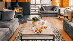 Een gezellige woonkamer met een open haard, grijze banken, een salontafel met eten en een groot raam.