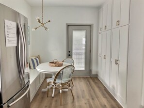 Une cuisine de taille réduite avec une table blanche, des chaises en osier, un réfrigérateur en acier inoxydable et des placards blancs.